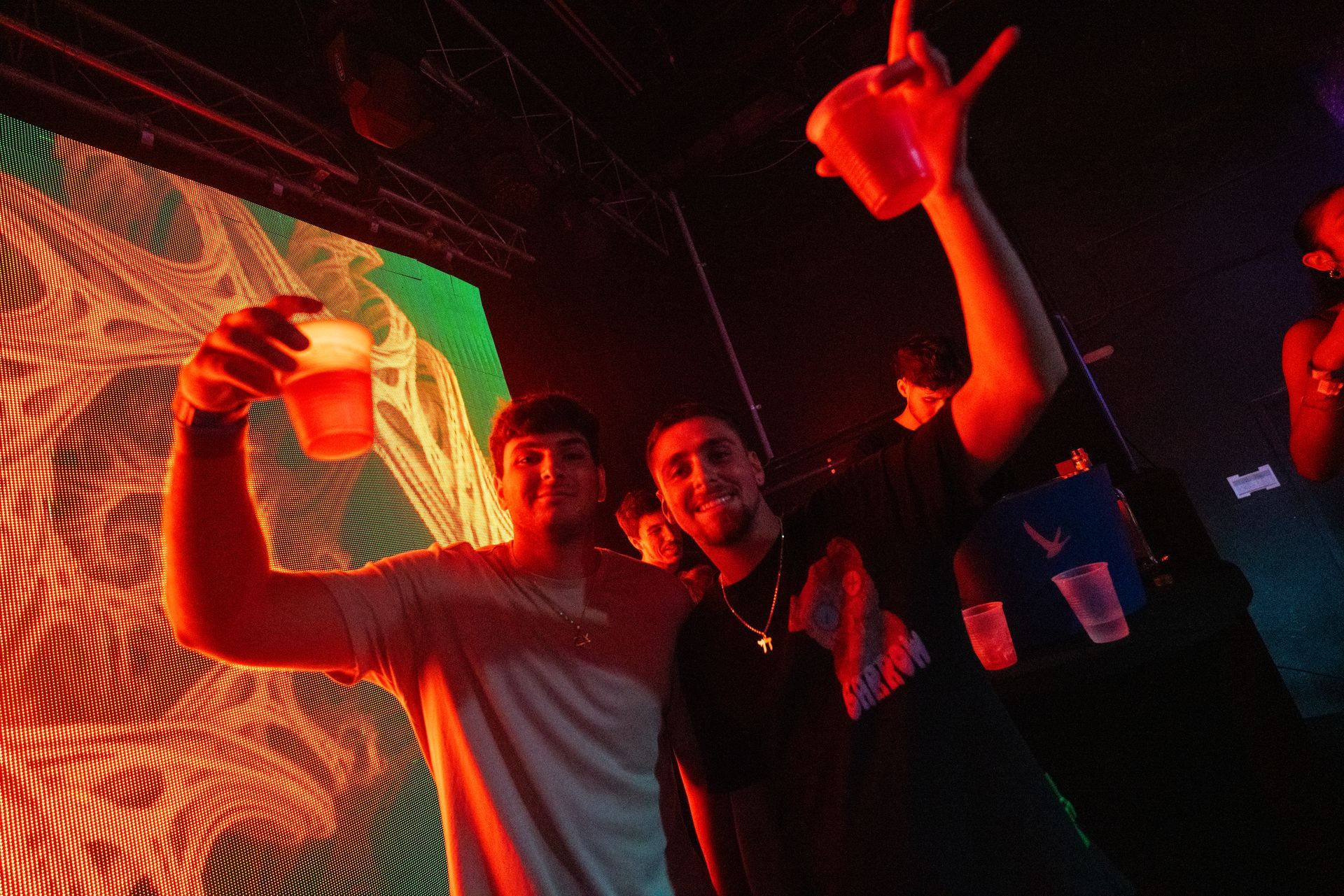 Two men are standing next to each other in a dark room holding cups in their hands.