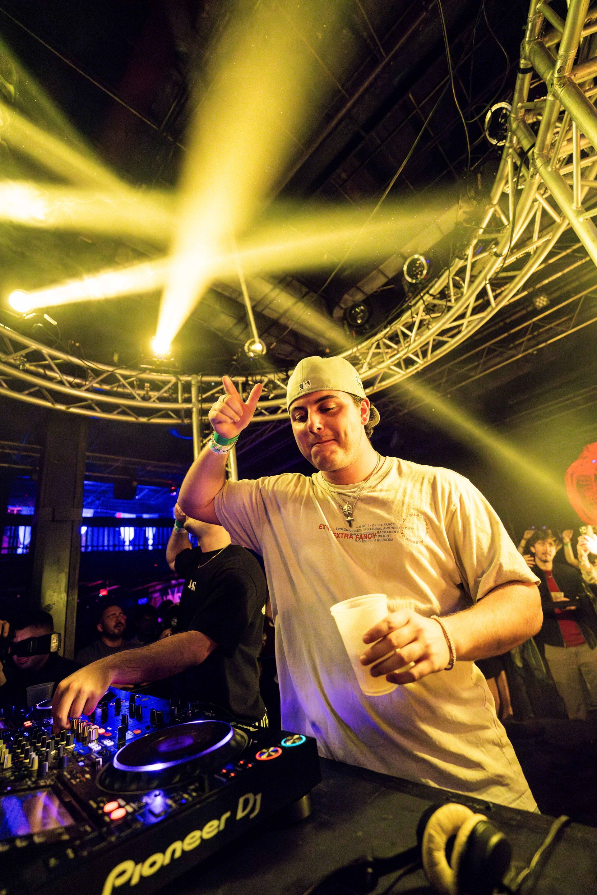 A man is playing a dj set in a club while holding a cup.