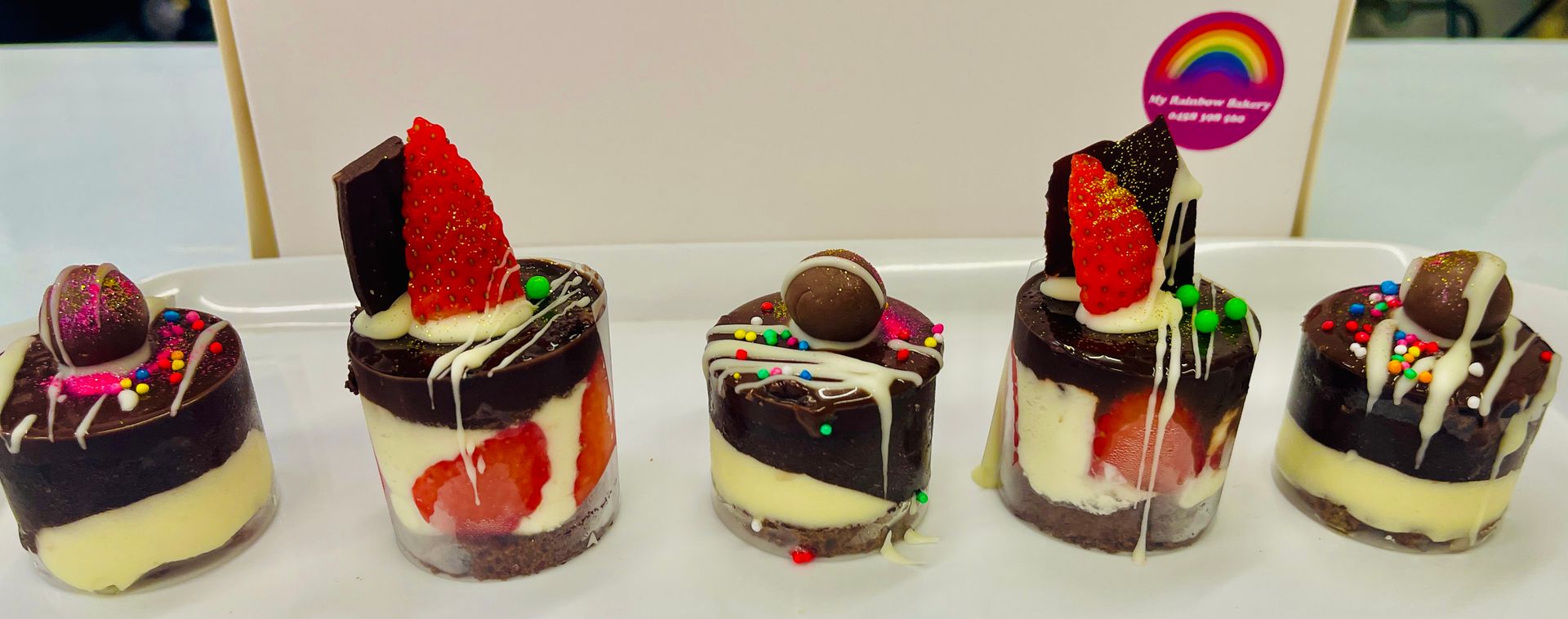 Five mini desserts on a white plate; chocolate, strawberry and a candy ball. White icing drizzle and sprinkles — My Rainbow Bakery, Cafe & Catering in Cannonvale, QLD