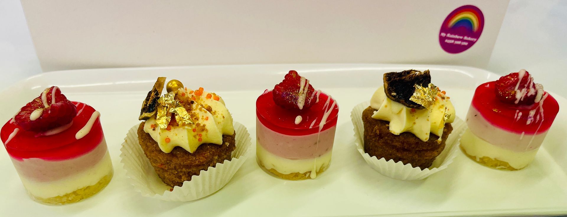 Five colorful mini cakes on a white platter. Each has a different topping and design, in front of a white box — My Rainbow Bakery, Cafe & Catering in Cannonvale, QLD