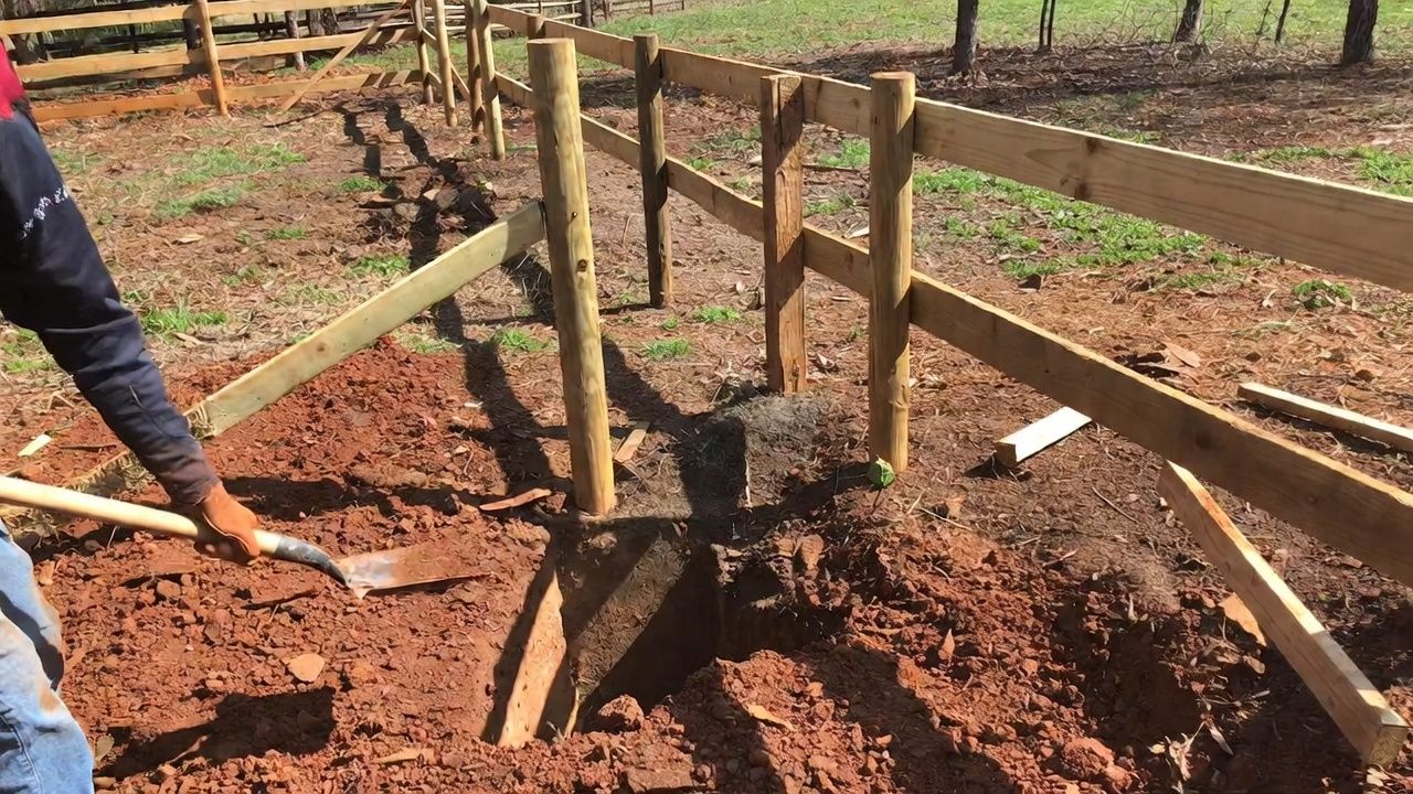 Person shoveling dirt around a wooden fence post, outdoors in a yard.