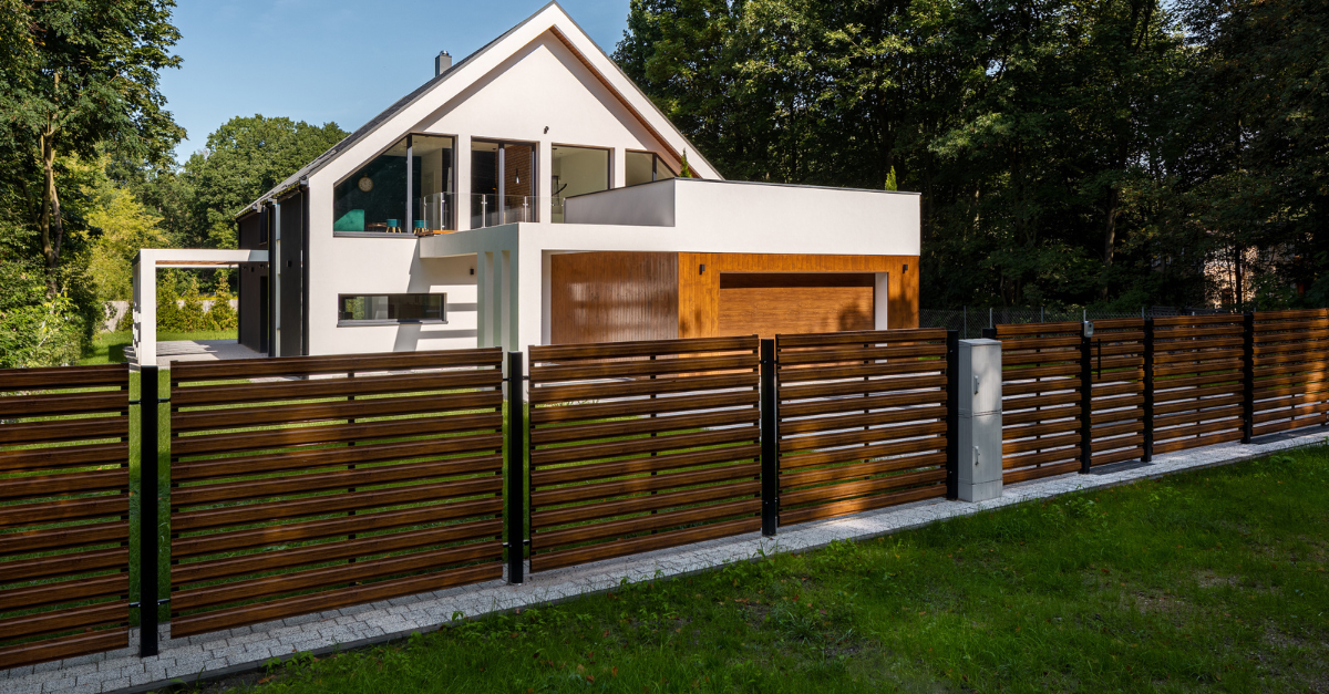 Modern house behind a woven wooden fence; green grass and trees in the background.