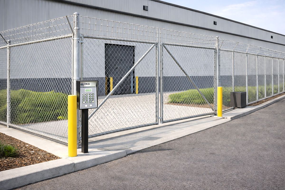 Chain link fence with barbed wire surrounding a building and gated entrance with access panel. Yellow bollards.