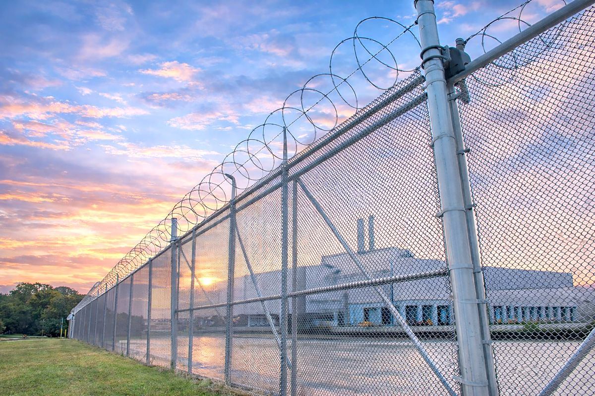 A chain-link fence topped with barbed wire, guarding a building against a sunset sky.