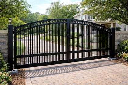 Black metal driveway gates with arched top, brick pillars, leading to a house.
