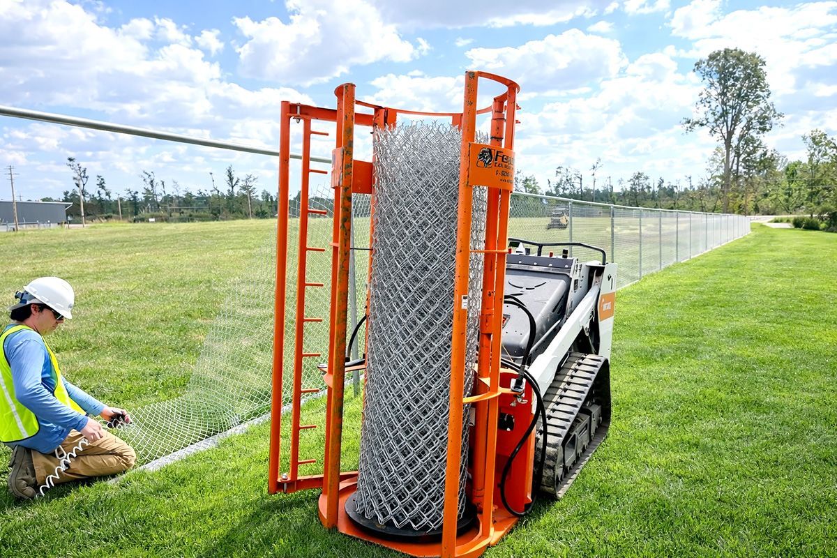 Orange machine with chain link fencing unrolls along a grassy fence line, a worker kneels adjusting the fence.
