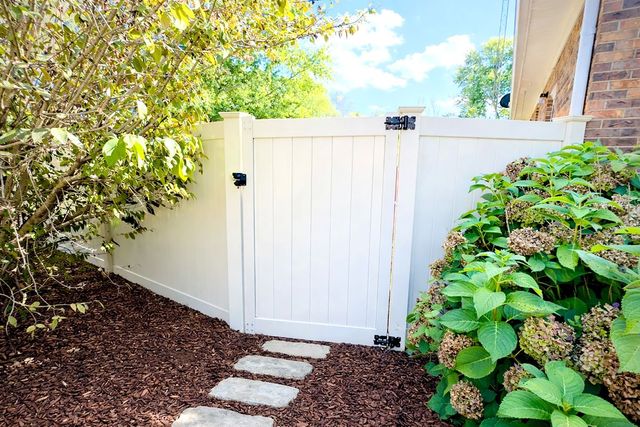 White picket gate in a garden, with stone path, brown mulch, and green foliage.