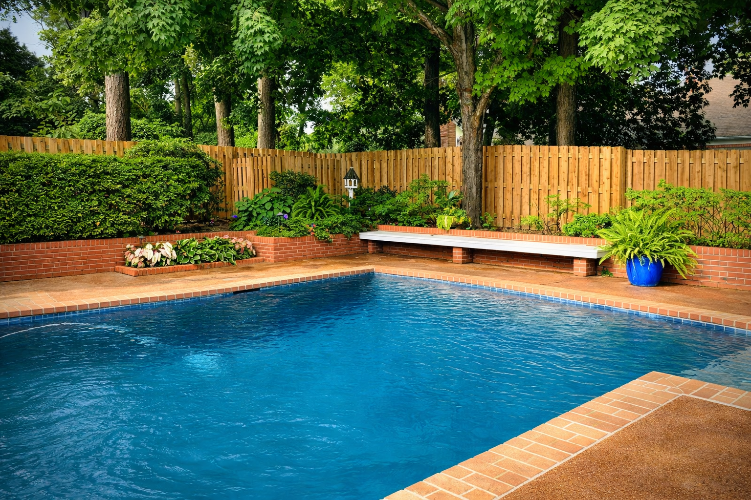Outdoor pool with blue water surrounded by a brick patio, wooden fence, greenery, and a bench.