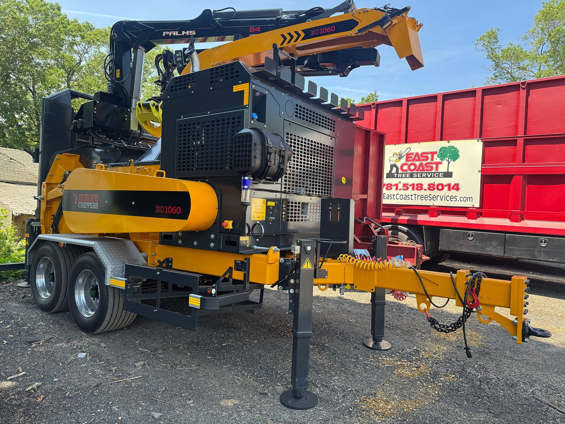 Yellow and black wood chipper machine with crane arm, parked next to a red dumpster.