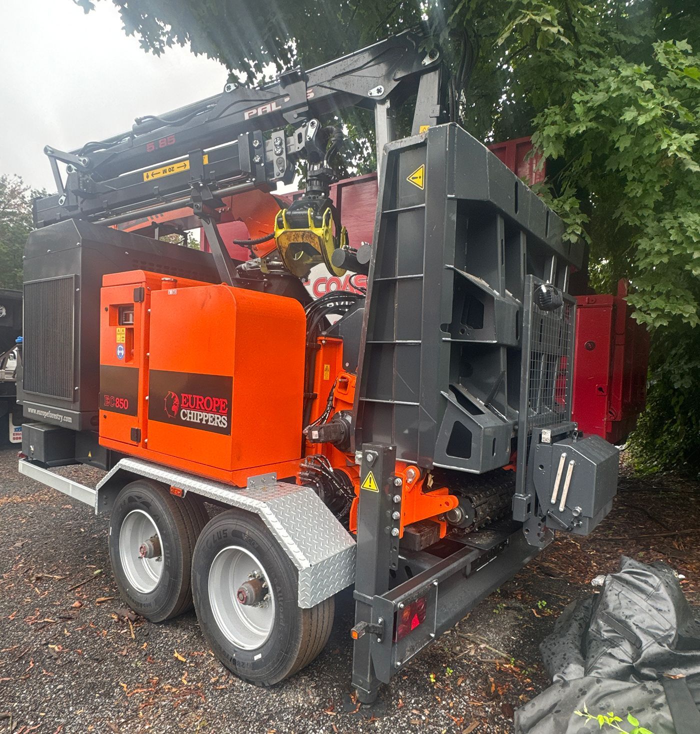 Orange and black wood chipper on a trailer, outdoors. The machine has a crane arm, multiple chutes, and a diesel engine.