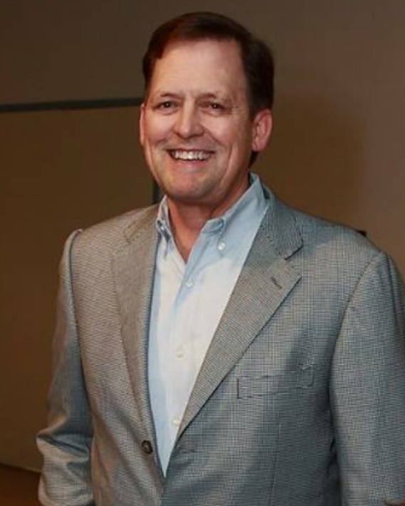Man in cowboy hat and brown blazer, leaning against a wall, with guitar.
