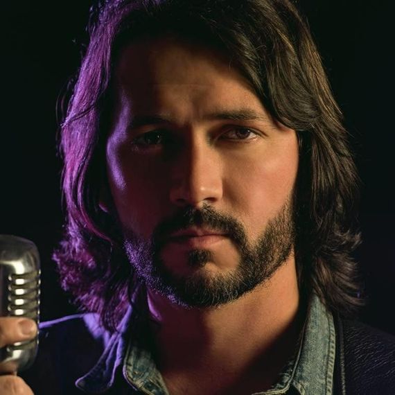 Man in cowboy hat and brown blazer, leaning against a wall, with guitar.
