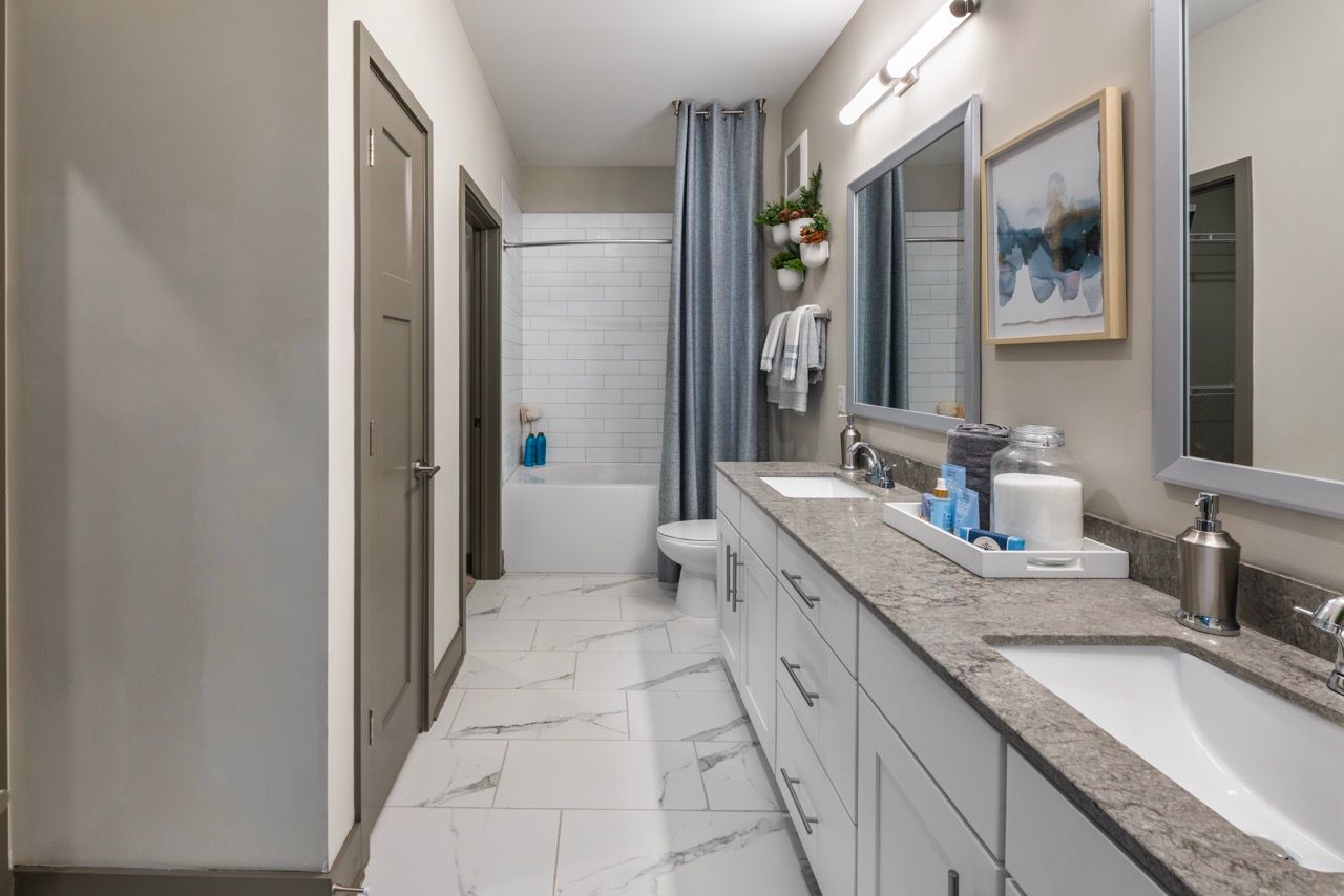Modern apartment bathroom with a double-sink vanity, granite countertop, and a white-tiled shower/tub.