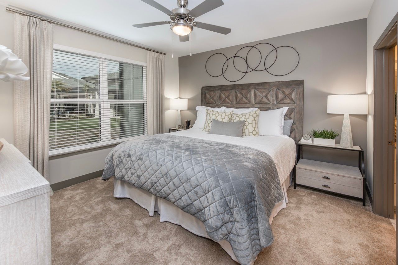 Bedroom in an apartment with a queen bed, gray quilted comforter, and a large window with curtains.