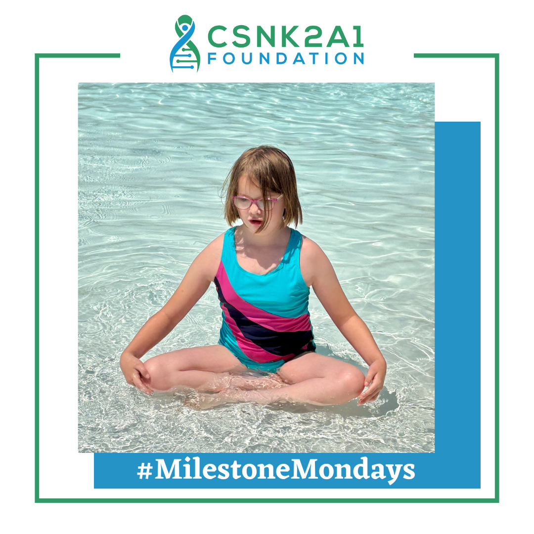 Girl in a teal and pink swimsuit sitting in shallow water, #MilestoneMondays.