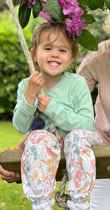 Smiling child on a swing, wearing green shirt and floral pants, outdoors under pink flowers.