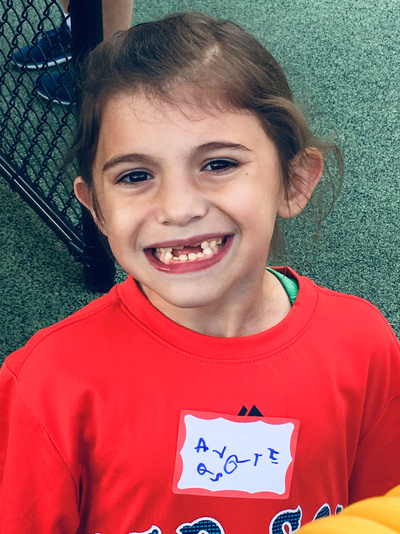 Girl in red shirt smiles, missing a tooth, with a name tag that says 