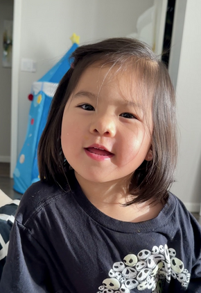 Smiling child with dark hair, wearing a black shirt, in a brightly lit room with a blue tent in the background.