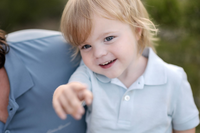 Blond toddler in a blue polo shirt pointing with a smile. Outdoors, leaning against someone in blue.