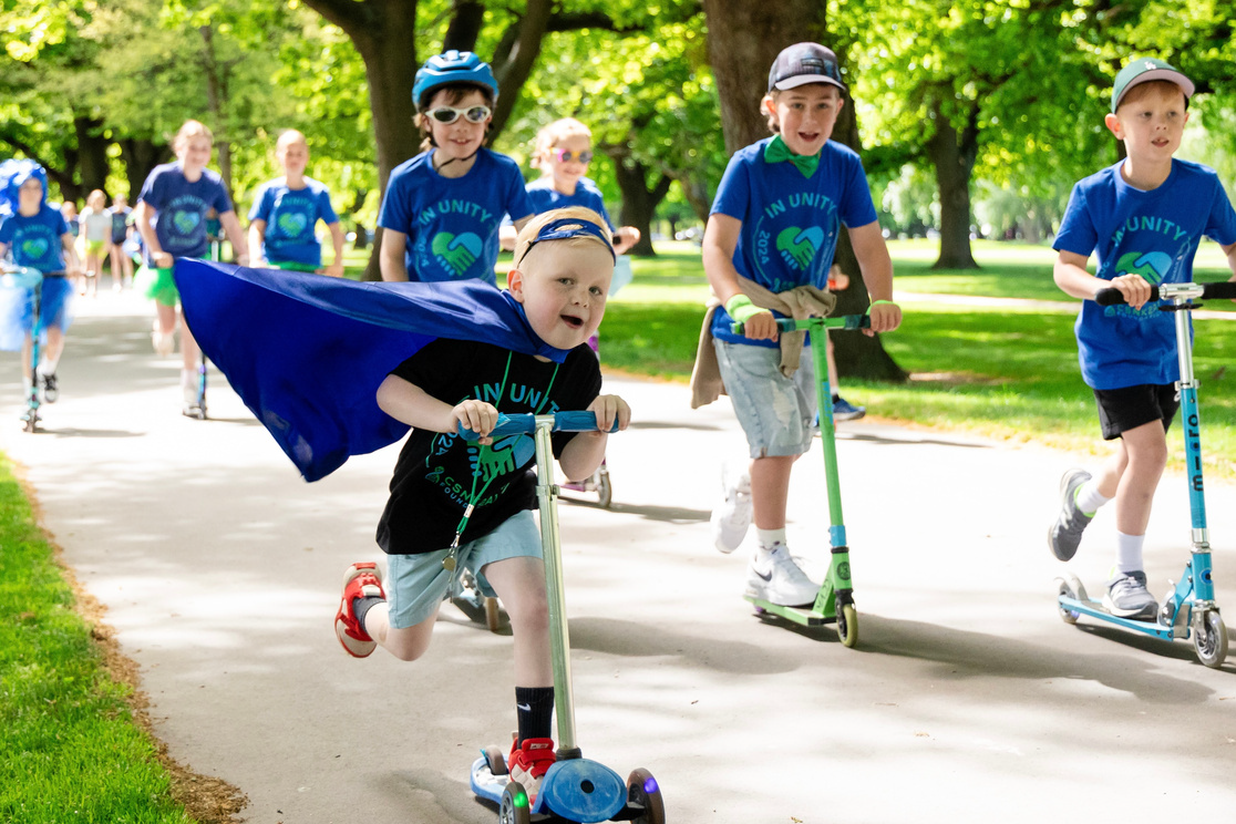 Children on scooters, one with a cape, racing down a paved path in a park; sunny day.