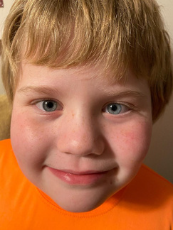 Blonde-haired child with blue eyes and rosy cheeks smiling in an orange shirt.