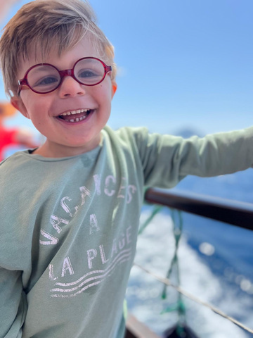 Boy with glasses smiles broadly on a boat, wearing a green sweatshirt with 
