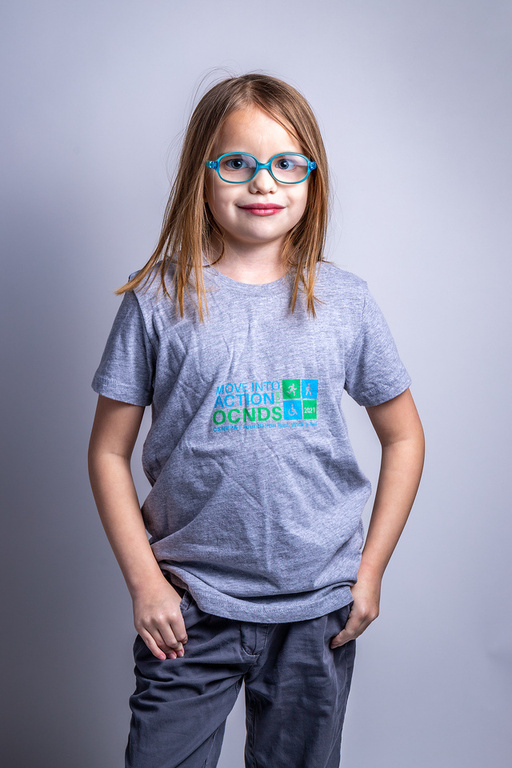 Girl wearing glasses and a gray t-shirt with a logo, standing with hands in pockets.