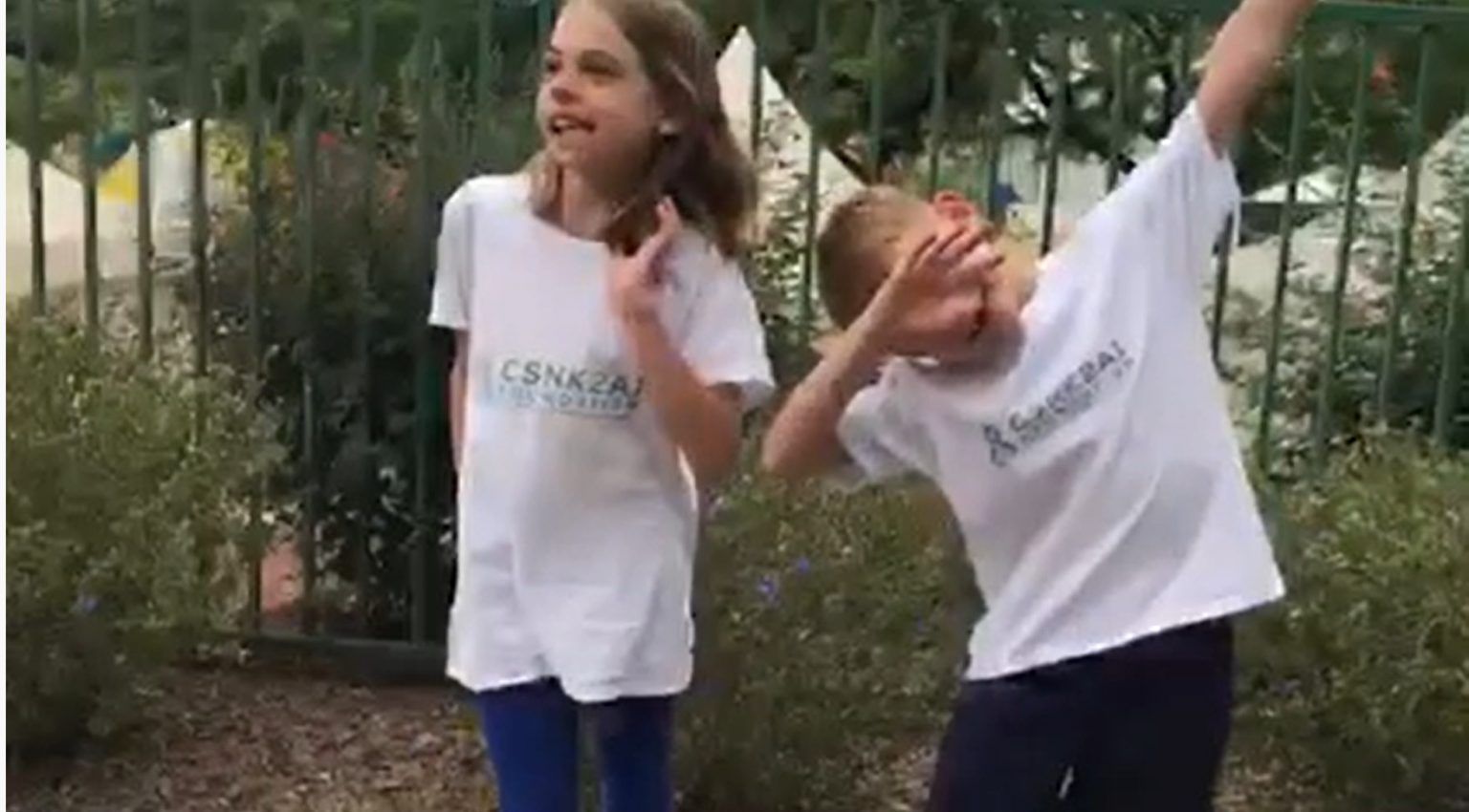 A boy and a girl are standing next to each other in front of a fence.