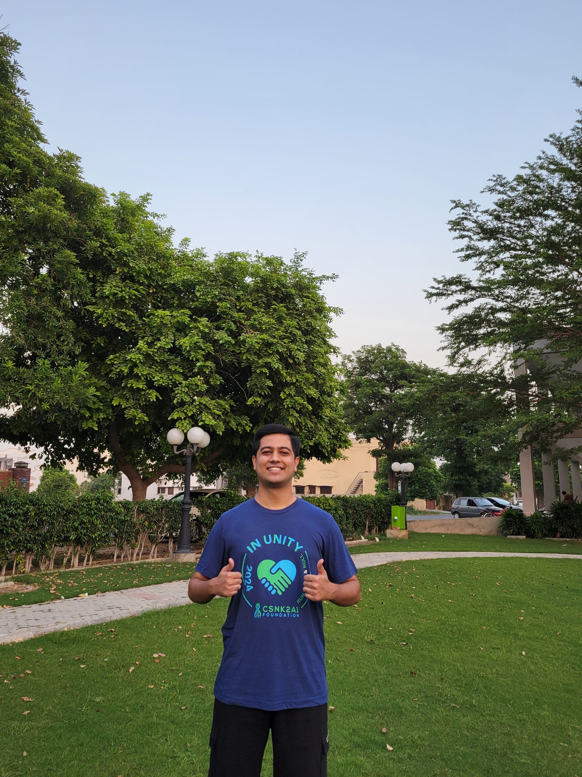 Person giving thumbs up in a park, wearing a blue t-shirt. Green trees and grass with a light blue sky in the background.