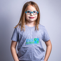 A little girl wearing glasses and a grey t-shirt is standing with her hands in her pockets.