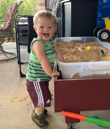 A little boy is standing next to a sandbox and smiling.