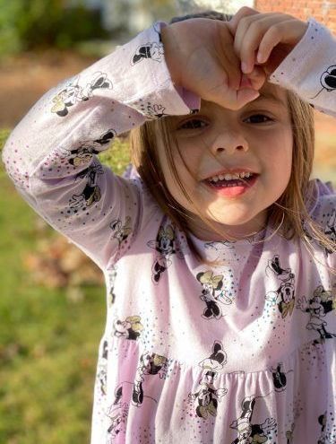 A little girl in a pink dress is covering her face with her hand.