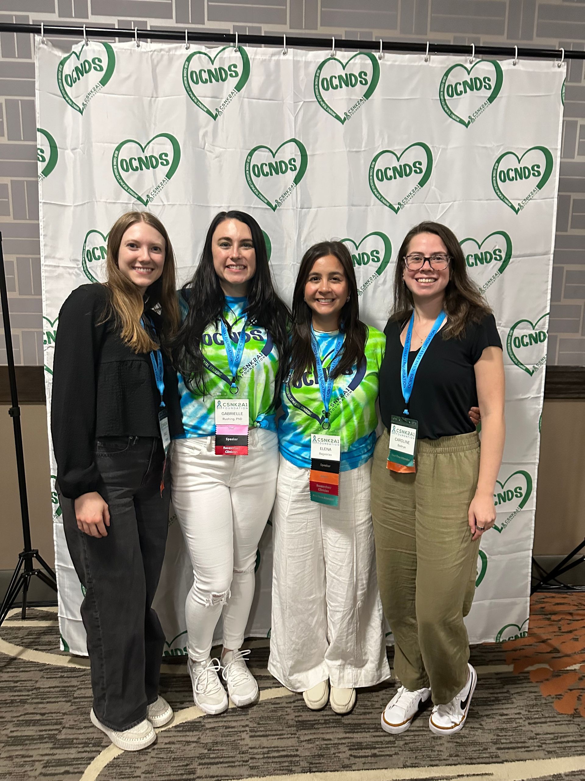 Four people smiling in front of a backdrop with a heart logo; two in tie-dye shirts, one in black, and one in green.