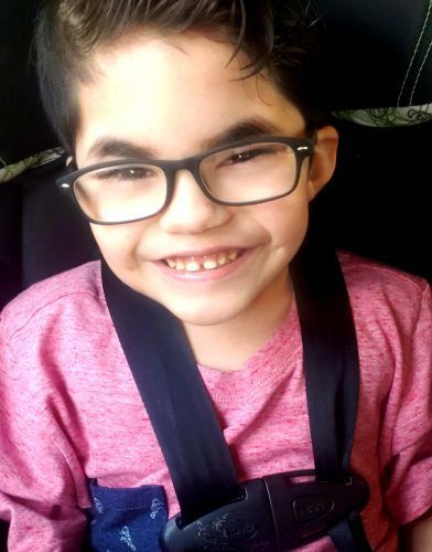 A young boy wearing glasses and a seat belt smiles for the camera.