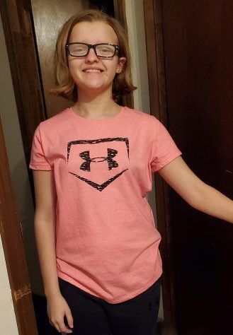 A young girl wearing glasses and a pink under armour shirt is standing in a hallway.