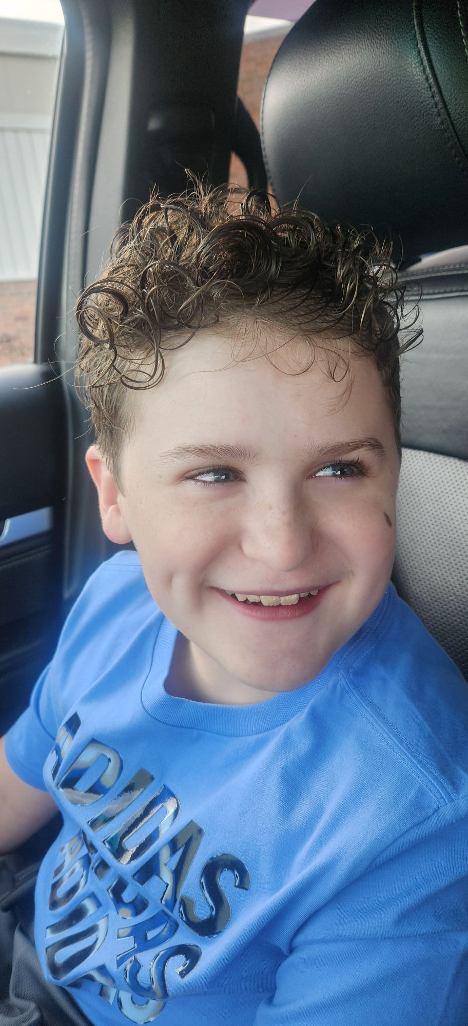 A smiling child in a blue shirt with curly hair. Seated inside a vehicle.