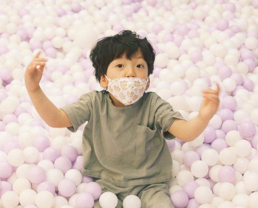 A young boy wearing a mask is sitting in a ball pit.
