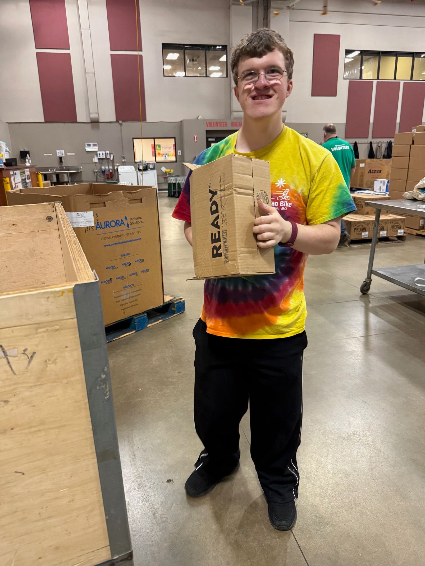 Young person in tie-dye shirt holding a cardboard box in a warehouse-like setting, smiling at the camera.