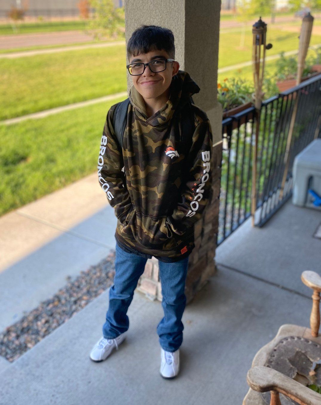 A young boy wearing a camo hoodie and glasses is standing on a porch.