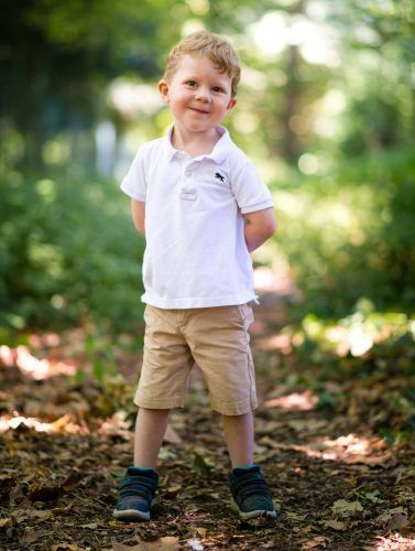 A young boy in a white shirt and khaki shorts is standing in the woods.