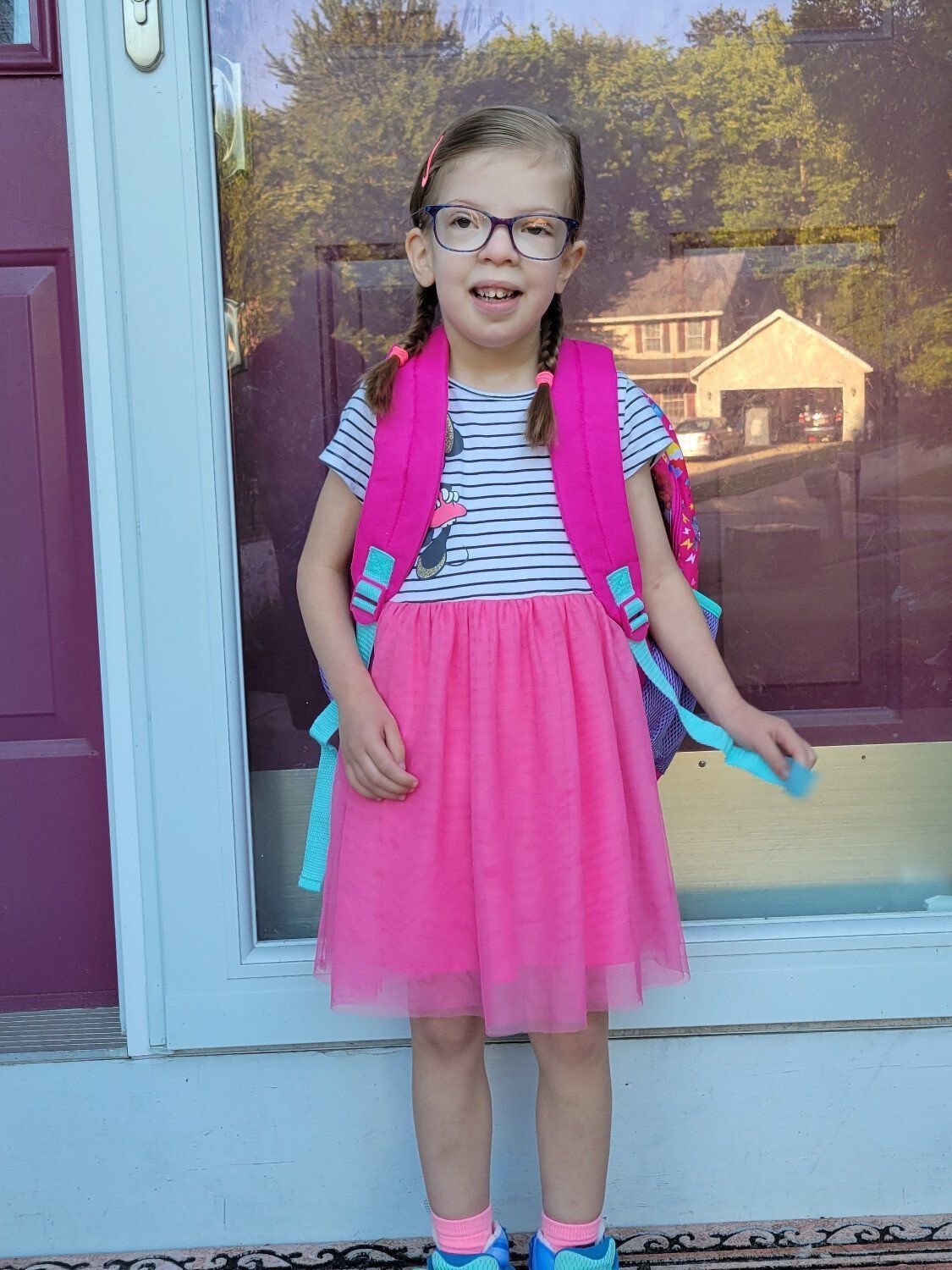 A little girl in a pink dress and glasses is standing in front of a door.