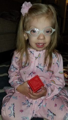 A little girl wearing glasses is sitting on the floor holding a red block.