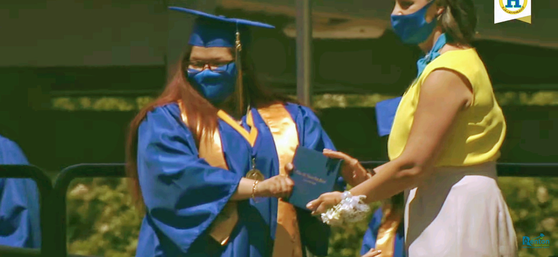 A woman wearing a mask is giving a diploma to a graduate.