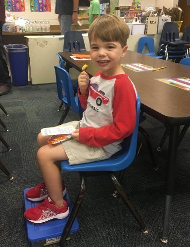 A young boy is sitting in a blue chair in a classroom eating a snack.