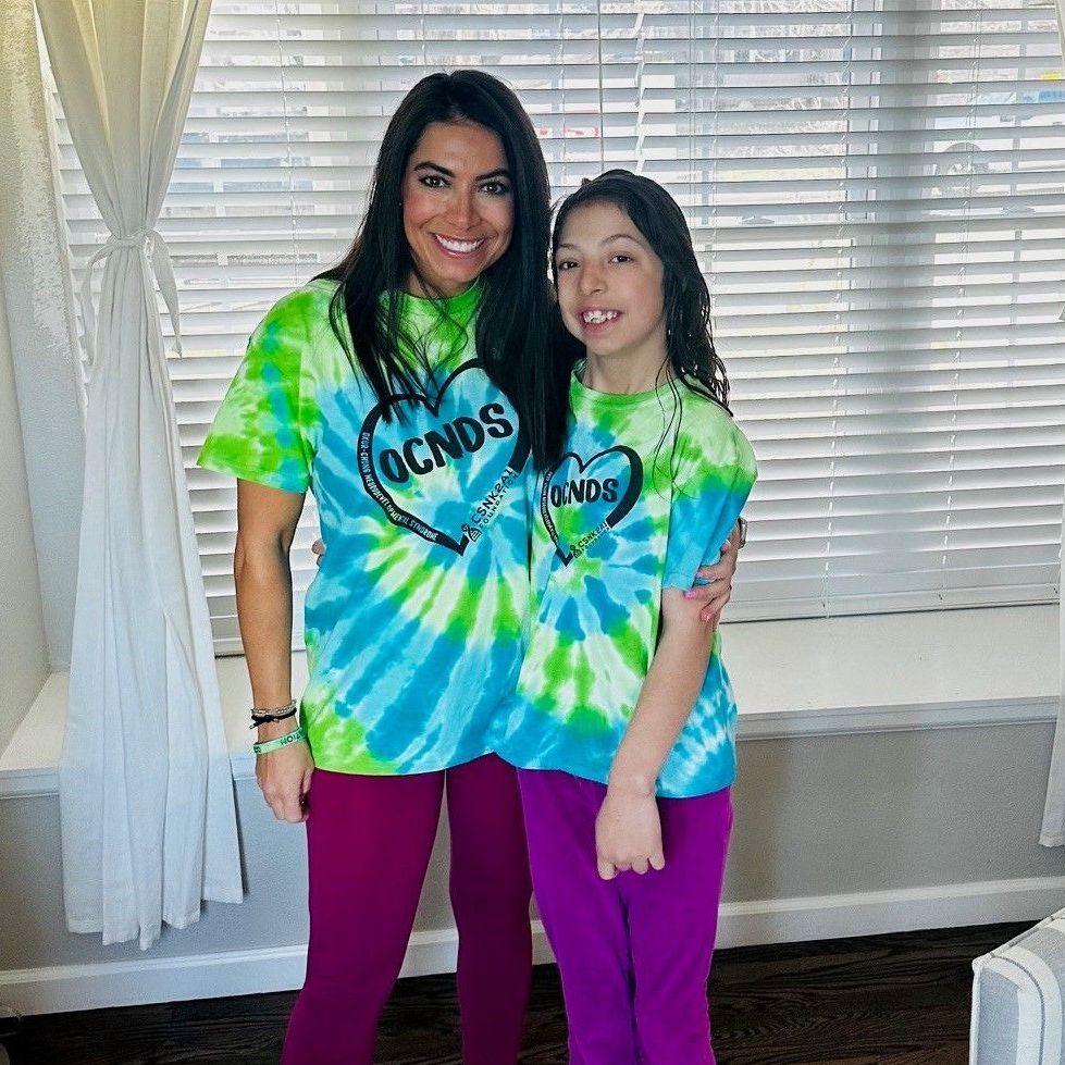 A woman and a girl are standing next to each other in a living room wearing tie dye shirts.