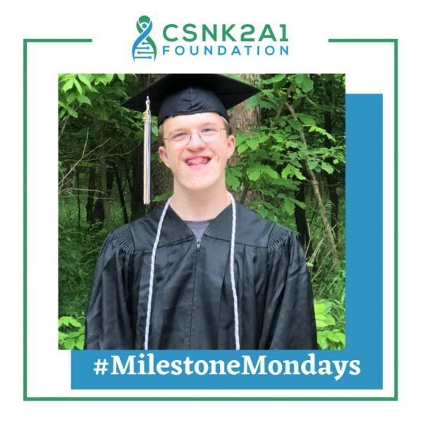 A young man in a graduation cap and gown stands in front of trees