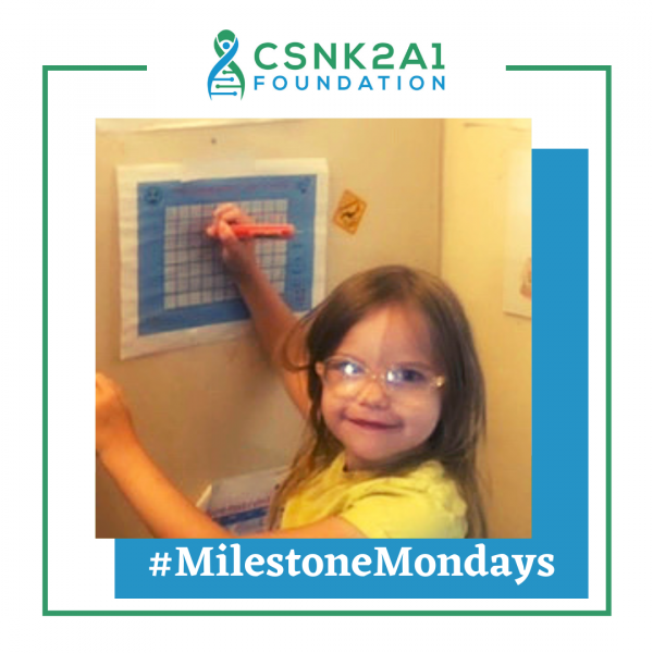 A little girl wearing glasses is writing on a board with the words milestone mondays below her