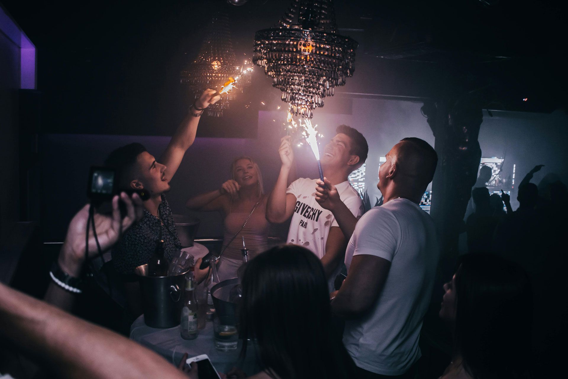 A dimly lit nightclub scene with a group of friends celebrating around a table with a glowing, sparkler-topped chandelier.