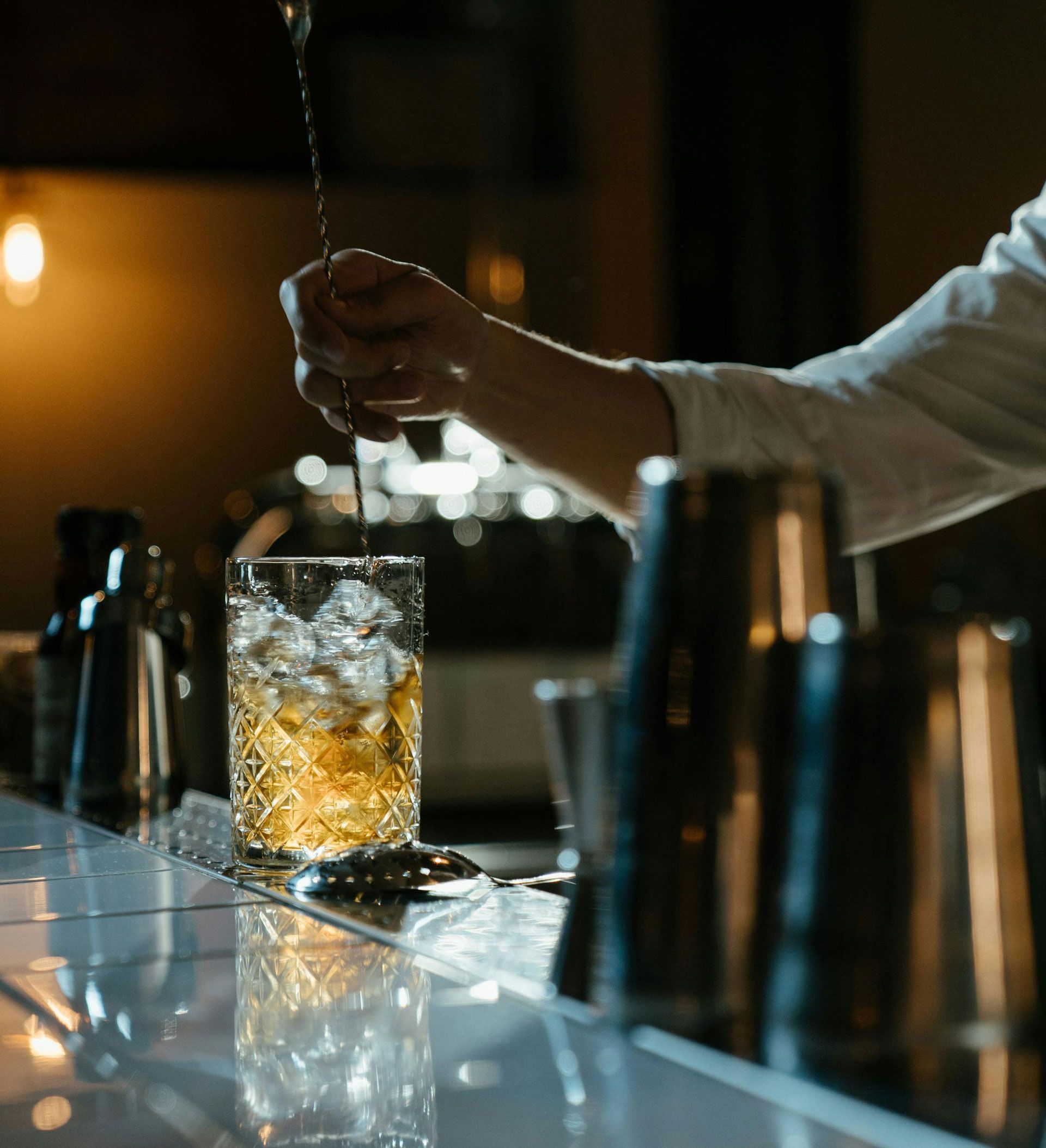 A bartender stirs an amber cocktail in a patterned glass filled with ice, set on a dark, reflective bar counter.
