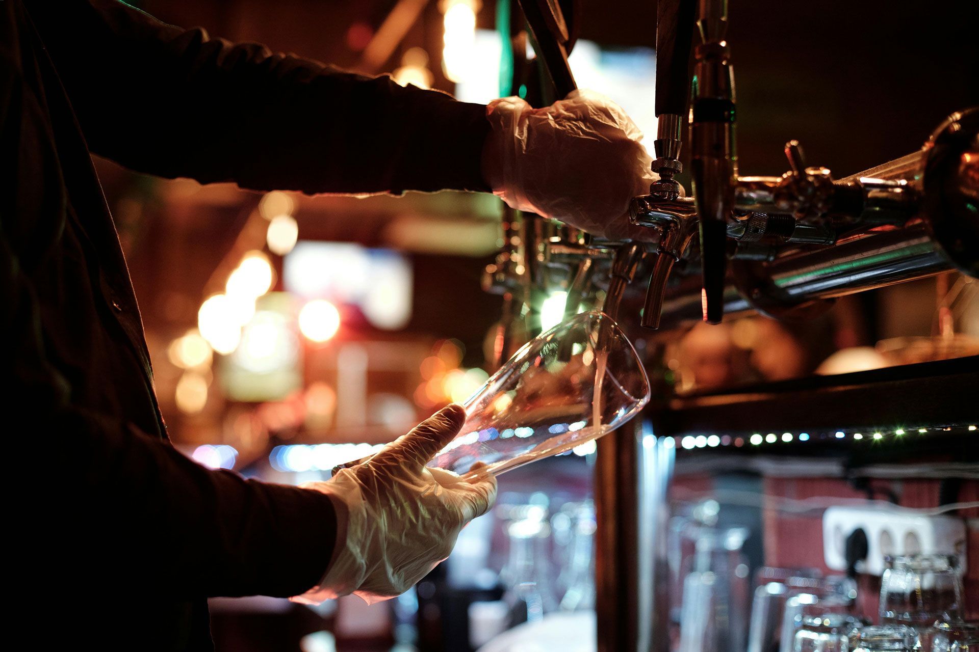 A person wearing disposable gloves pours draft beer into a glass behind a dimly lit bar.